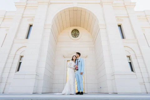 Hombre con traje celeste y mujer con vestido blanco aparecen de pie a distancia bajo un gran arco del edificio.