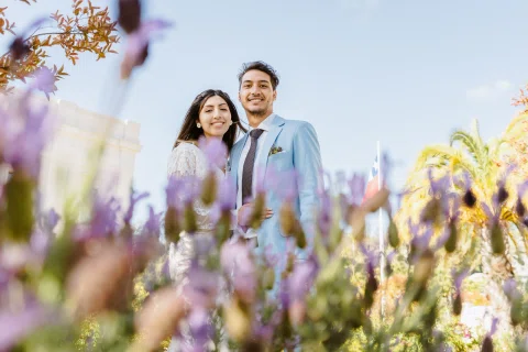 Hombre con traje celeste y mujer con vestido blanco aparecen juntos detrás de flores lilas desenfocadas.