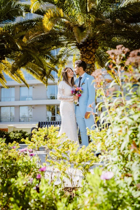 Hombre con traje celeste y mujer con vestido blanco aparecen juntos entre flores y palmeras.