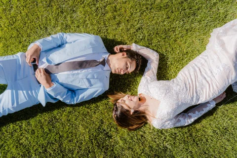 Hombre con traje celeste y mujer con vestido blanco están recostados sobre el césped, fotografiados desde arriba.