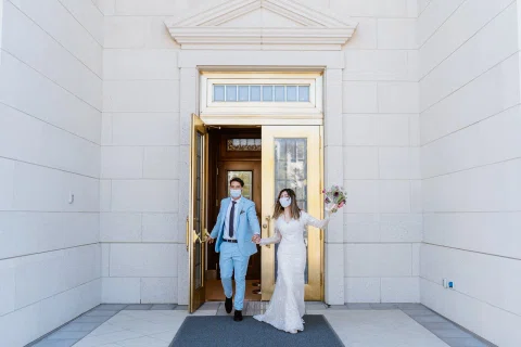 Hombre con traje celeste y mujer con vestido blanco salen juntos por la puerta de un edificio blanco.