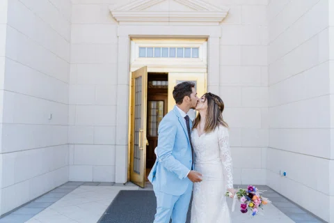 Hombre con traje celeste y mujer con vestido blanco se besan frente a la puerta de un edificio blanco.
