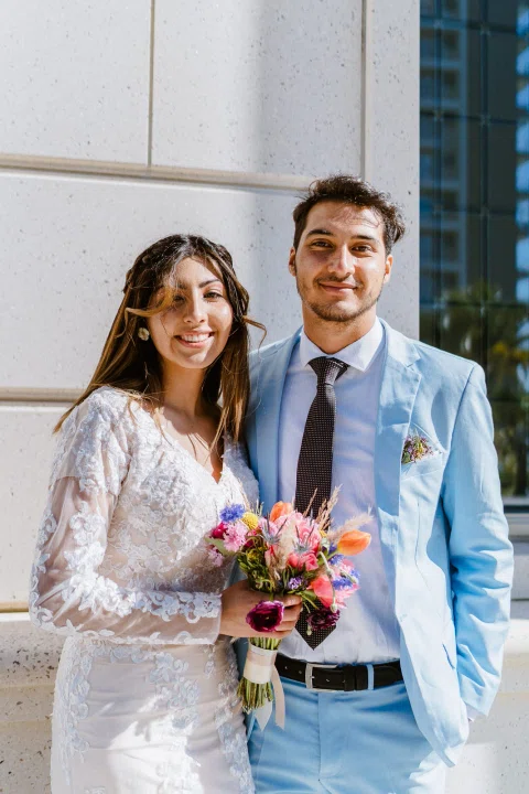 Hombre con traje celeste y mujer con vestido blanco sonríen juntos mientras ella sostiene un ramo.