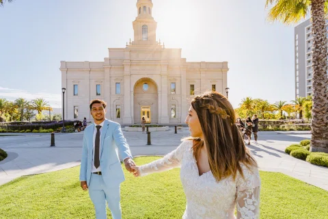 Hombre con traje celeste y mujer con vestido blanco están tomados de la mano frente a un edificio blanco.
