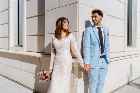 Hombre con traje celeste y mujer con vestido blanco están tomados de la mano junto a un muro claro.