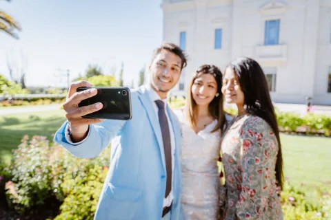 Hombre con traje celeste toma una selfie junto a dos mujeres en un jardín.