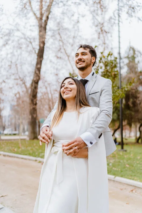 Hombre con traje claro rodea con los brazos a una mujer con vestido blanco desde atrás; ambos sonríen en un parque.