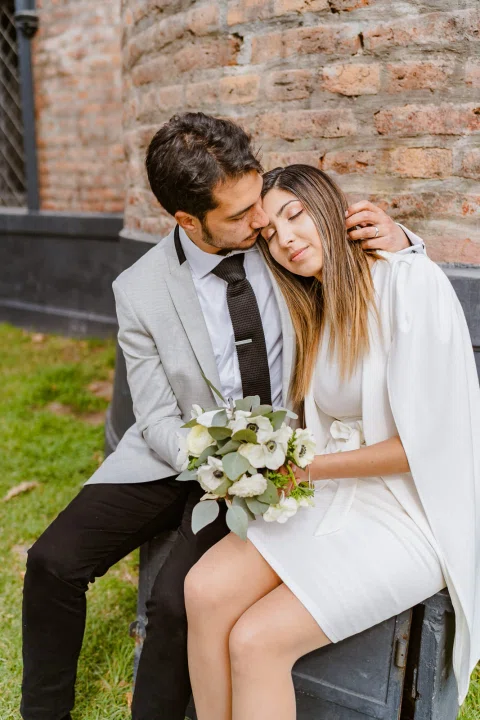 Hombre con traje claro besa la cabeza de una mujer con vestido blanco mientras ambos están sentados junto a un muro de ladrillo; ella sostiene un ramo.