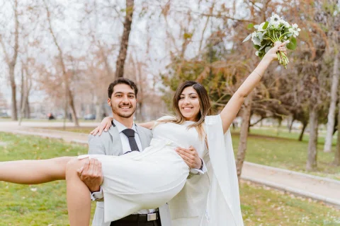 Hombre con traje claro carga en brazos a una mujer con vestido blanco en un parque; ella levanta un ramo.