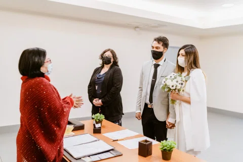 Hombre con traje claro y mujer con vestido blanco permanecen frente a una mujer vestida de rojo junto a un escritorio.