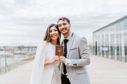 Hombre con traje claro y mujer con vestido blanco posan juntos sosteniendo una libreta en una terraza.