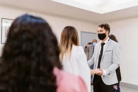 Hombre con traje claro y mascarilla sostiene la mano de una mujer con vestido blanco mientras otras personas permanecen alrededor.