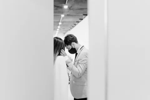 Hombre con traje claro y mascarilla inclina la cabeza mientras toca el cabello de una mujer, en una fotografía en blanco y negro.