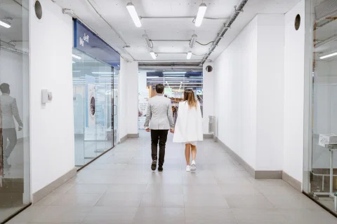 Hombre con traje claro y mujer con vestido blanco caminan de espaldas por un pasillo largo y luminoso.