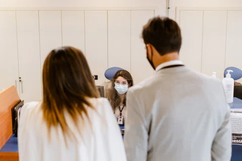 Hombre con traje claro y mujer con vestido blanco aparecen de espaldas frente a un mostrador dentro de una oficina.
