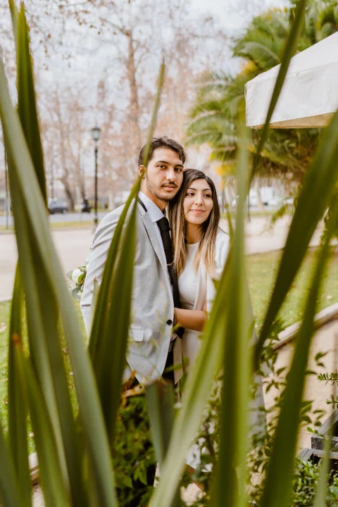 Hombre con traje claro y mujer con vestido blanco posan juntos detrás de hojas de palmera.