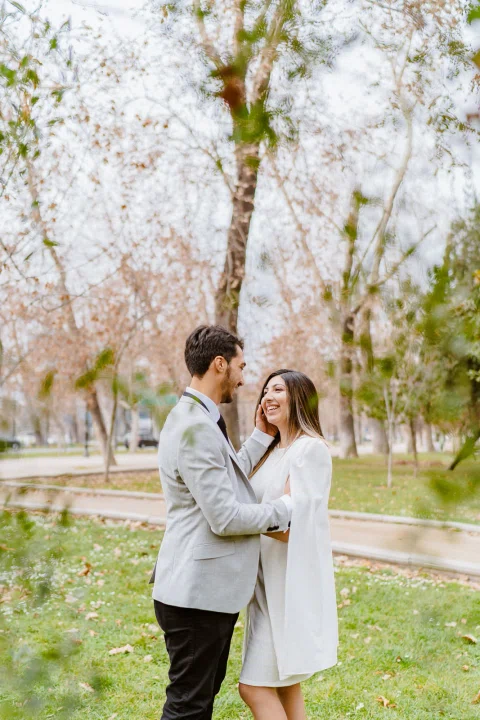 Hombre con traje claro y mujer con vestido blanco se miran sonriendo frente a frente en un parque.