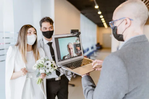 Hombre con traje claro y mujer con vestido blanco observan un computador portátil sostenido por otro hombre; en la pantalla aparece un rostro.