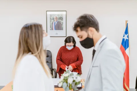 Hombre con traje claro y mujer con vestido blanco permanecen frente a una mujer vestida de rojo junto a una bandera de Chile.