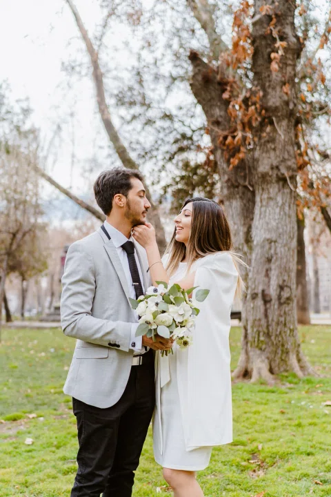 Hombre con traje claro y mujer con vestido blanco permanecen junto a un árbol; ella sostiene un ramo y toca el rostro de él.