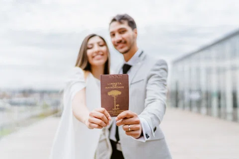 Hombre con traje claro y mujer con vestido blanco sonríen y sostienen una libreta hacia la cámara en un espacio exterior.
