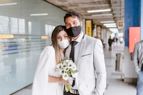 Hombre con traje claro y mujer con vestido blanco posan juntos en un pasillo interior; ella sostiene un ramo y ambos usan mascarilla.