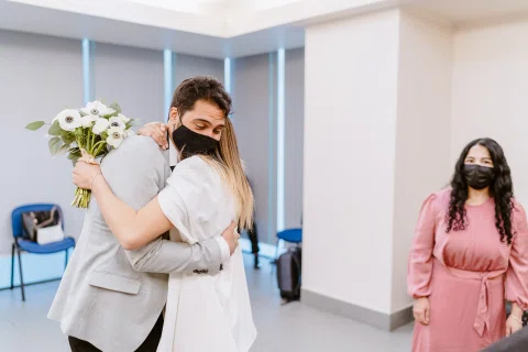 Hombre con traje claro y mujer con vestido blanco se abrazan mientras una mujer vestida de rosado permanece a un costado.