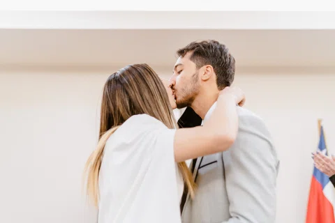Hombre con traje claro y mujer con vestido blanco se besan de perfil dentro de una sala.