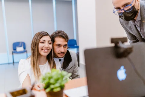 Hombre con traje claro y mujer con vestido blanco permanecen sentados junto a un computador portátil mientras miran la pantalla.