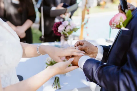 Hombre con traje azul coloca un anillo en la mano de una mujer con vestido blanco durante la ceremonia al aire libre.