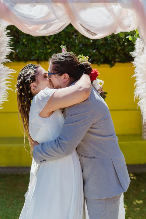 Hombre con traje gris abraza y besa a una mujer con vestido blanco bajo un arco decorado.