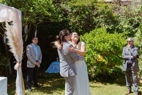 Hombre con traje gris abraza a una mujer con vestido blanco junto a un arco decorado.
