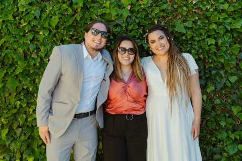 Hombre con traje gris, mujer con blusa coral y mujer con vestido blanco posan frente a un muro cubierto de hojas verdes.