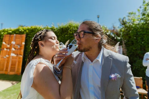 Hombre con traje gris y mujer con vestido blanco beben de copas con los brazos cruzados.