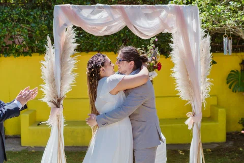 Hombre con traje gris y mujer con vestido blanco se besan bajo un arco decorado.