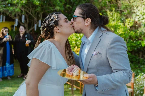 Hombre con traje gris y mujer con vestido blanco se besan mientras él sostiene un plato dorado con una porción de torta.