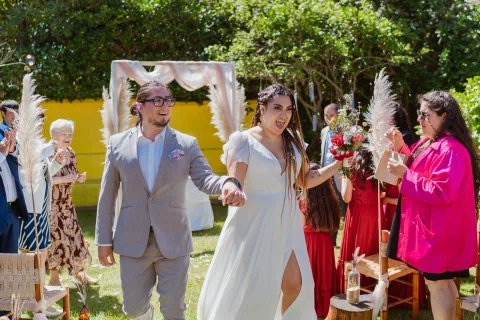 Hombre con traje gris y mujer con vestido blanco caminan de la mano entre personas en el jardín.