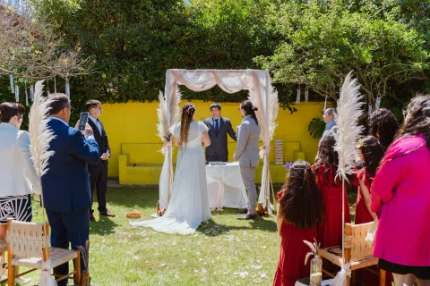 Hombre con traje gris y mujer con vestido blanco están frente a frente bajo un arco, con un tercer hombre detrás.