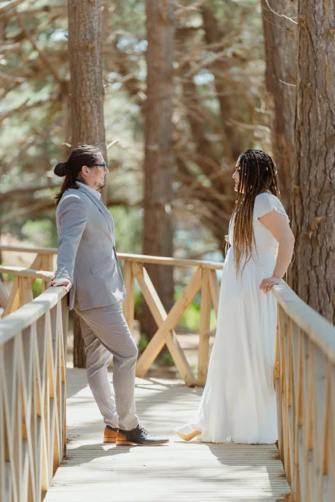 Hombre con traje gris y mujer con vestido blanco se miran junto a la baranda de una pasarela entre árboles.