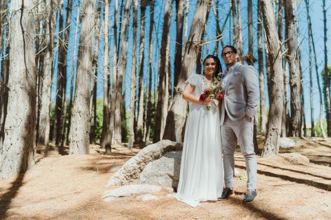 Hombre con traje gris y mujer con vestido blanco posan de cuerpo entero entre altos troncos de árboles.