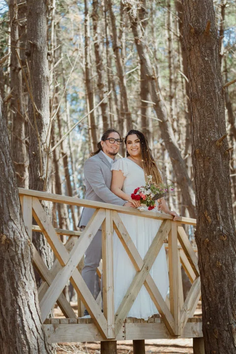 Hombre con traje gris y mujer con vestido blanco posan juntos junto a una baranda de madera entre árboles.