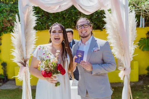 Hombre con traje gris y mujer con vestido blanco sonríen bajo un arco; ella sostiene un ramo y él un pequeño cuaderno morado.