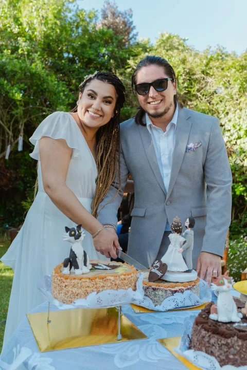 Hombre con traje gris y mujer con vestido blanco sonríen mientras cortan juntos una torta decorada.