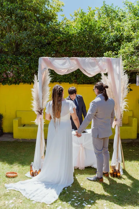 Hombre con traje gris y mujer con vestido blanco, vistos de espaldas, permanecen tomados de la mano frente a una mesa bajo un arco.