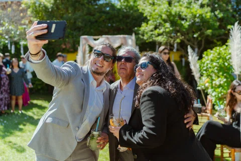 Hombre con traje gris sostiene un celular para una selfie junto a un hombre y una mujer con lentes de sol.