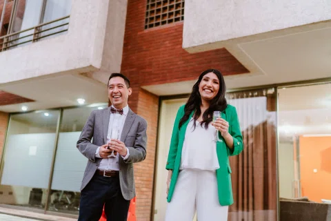 Hombre con traje gris sostiene una copa mientras ríe junto a una mujer con blazer verde en un patio.