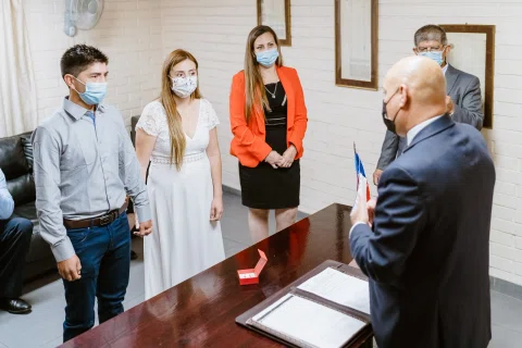 Hombre con traje y mascarilla habla frente a varias personas dentro de una sala.