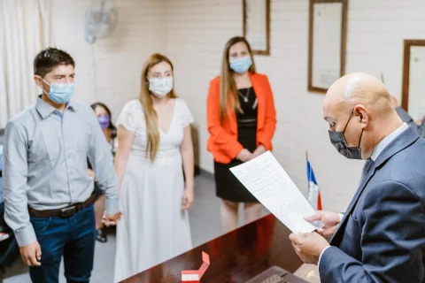 Hombre con traje y mascarilla lee un documento detrás de una mesa.