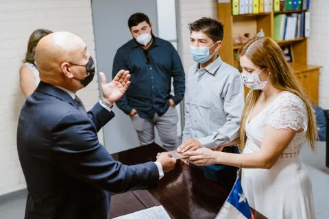 Hombre con traje y mascarilla sostiene la mano de una mujer con vestido blanco frente a un hombre con camisa celeste.