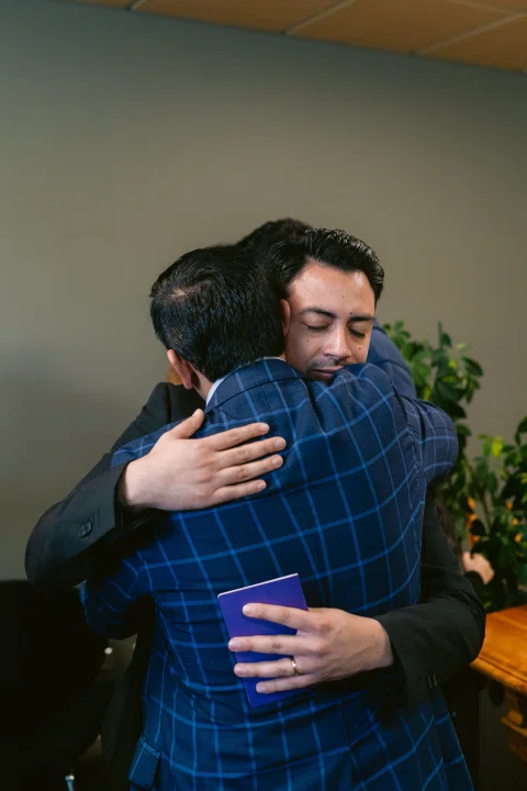 Hombre de traje negro abraza a un hombre con camisa a cuadros.